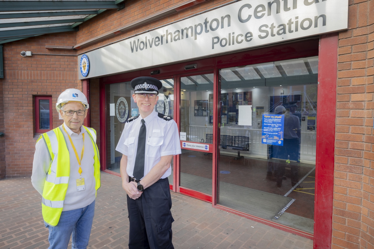 Two men stand in front of a police station. One man is in a high vis vest and construction hard hat and the other is a police uniform.
