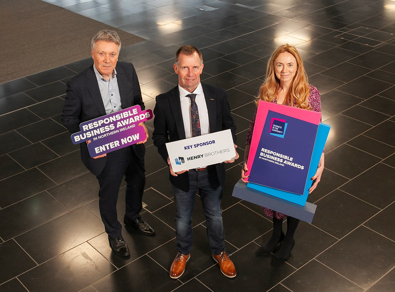 Kieran Harding, Managing Director of BITC NI, Ian Henry, CR Director at Henry Brothers, and Vicky Davies, Chair, Business in the Community, at the launch of the Responsible Business Awards in Northern Ireland. Three people stand in a line with props, announcing the open of Business in the Community's Responsible Business Awards in Northern Ireland. The first is a man in a black suit holding a shaped board that reads "Responsible Business Awards in Northern Ireland. Enter now". The second man wears jeans and a blazer, with a rectangle prop that reads "Key Sponsor: Henry Brothers". The third is a lady who holds a trophy shaped prop that reads "Responsible Business Awards in Northern Ireland".